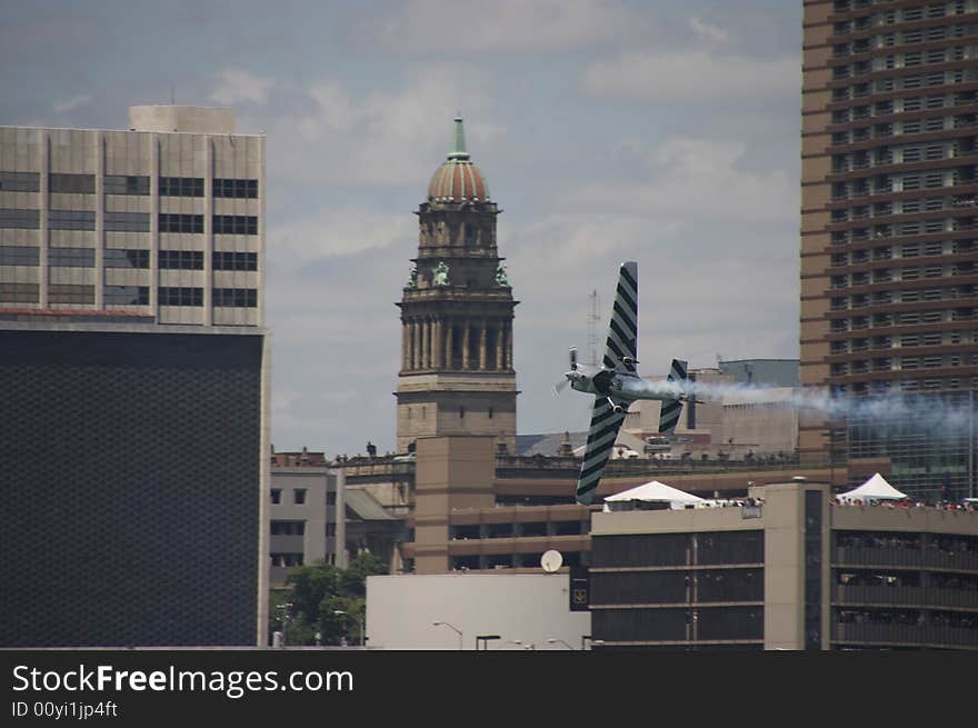 Air race plane doing stunt flying through the city with a crowd of people watching in the background.