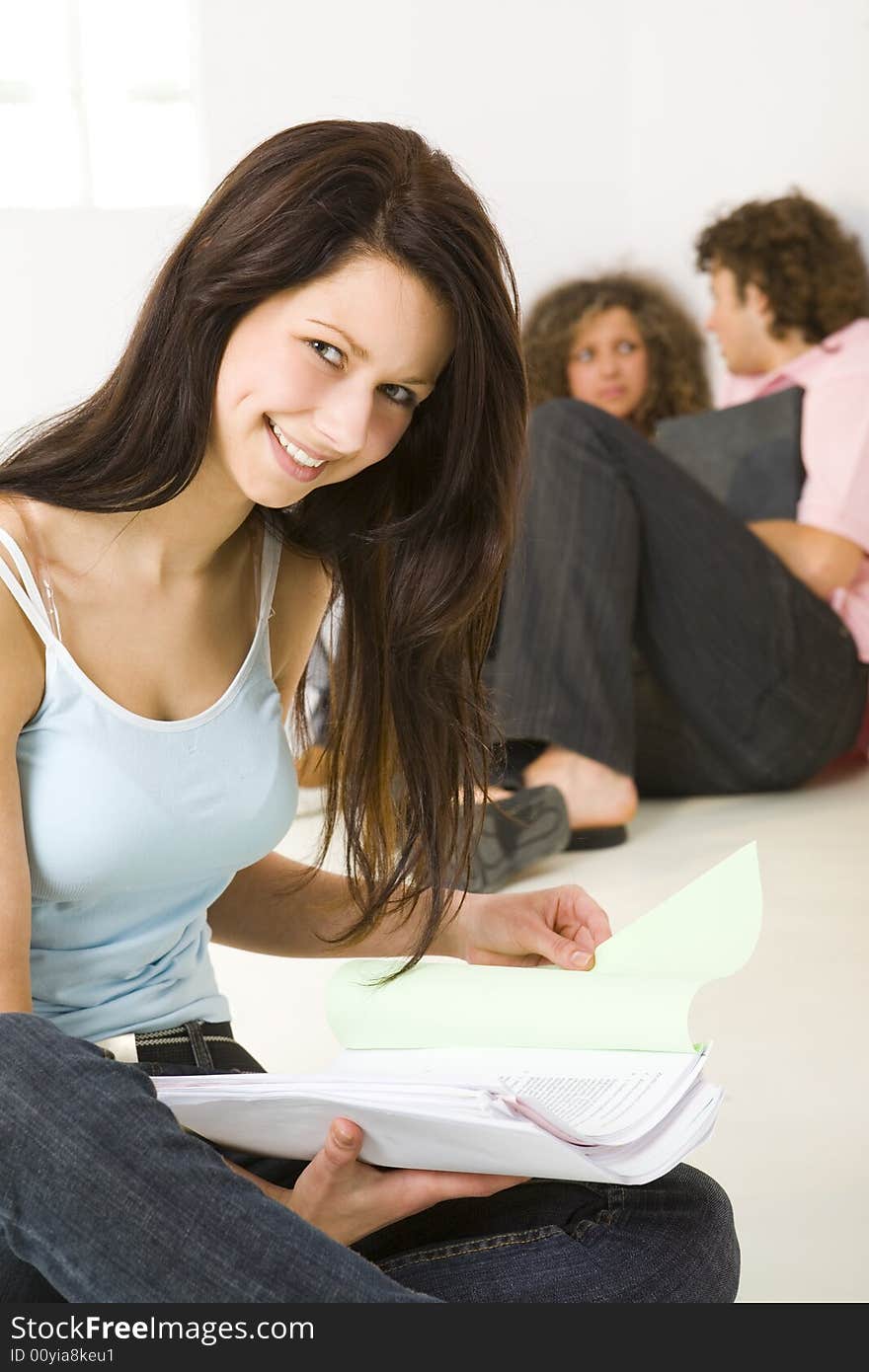 Three schoolmate holding a notebooks and sitting on the floor. A boy talking with girl in pink shirt. A girl in blue shirt smiling and looking at camera. Focused on girl in blue shirt. Three schoolmate holding a notebooks and sitting on the floor. A boy talking with girl in pink shirt. A girl in blue shirt smiling and looking at camera. Focused on girl in blue shirt.