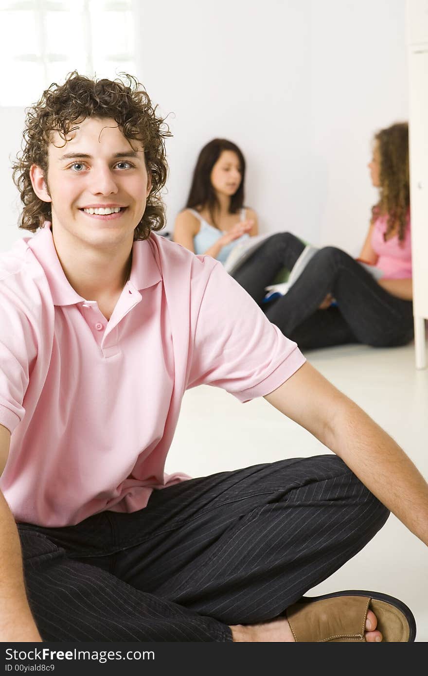 Three friends sitting on the floor. A girls talking with themselves. A boy smiling and looking at camera. Focused on a boy. Three friends sitting on the floor. A girls talking with themselves. A boy smiling and looking at camera. Focused on a boy.