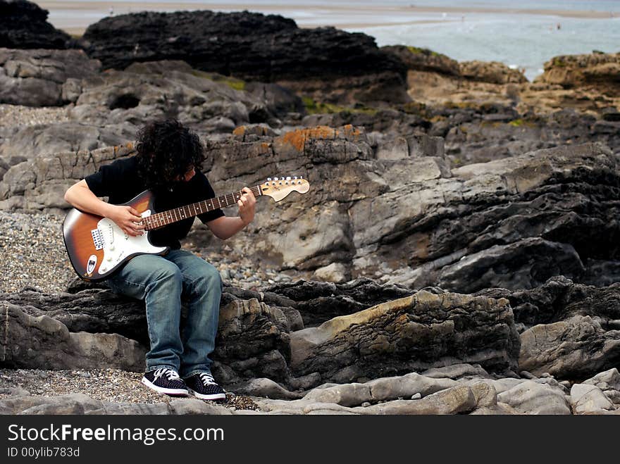 A teenage boy playing the guitar on the rocks near the beach. A teenage boy playing the guitar on the rocks near the beach.