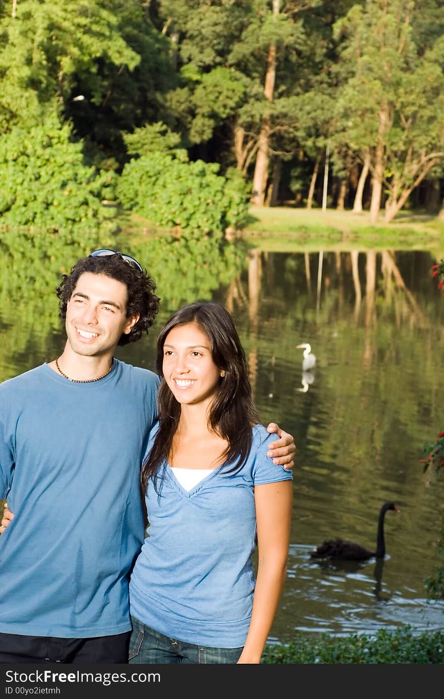 Happy, smiling couple embrace as they stand in front of a wooded pond with swans in it. Vertically framed photograph. Happy, smiling couple embrace as they stand in front of a wooded pond with swans in it. Vertically framed photograph.