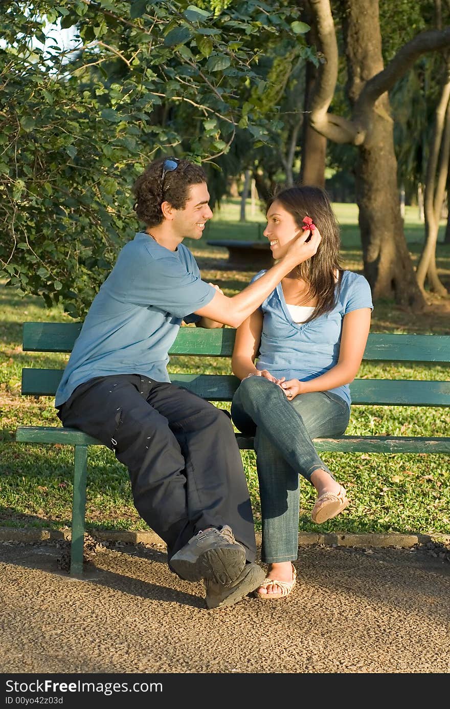 Happy couple on a park bench smile at each other as he places a red flower behind her ear. Vertically framed photograph. Happy couple on a park bench smile at each other as he places a red flower behind her ear. Vertically framed photograph.