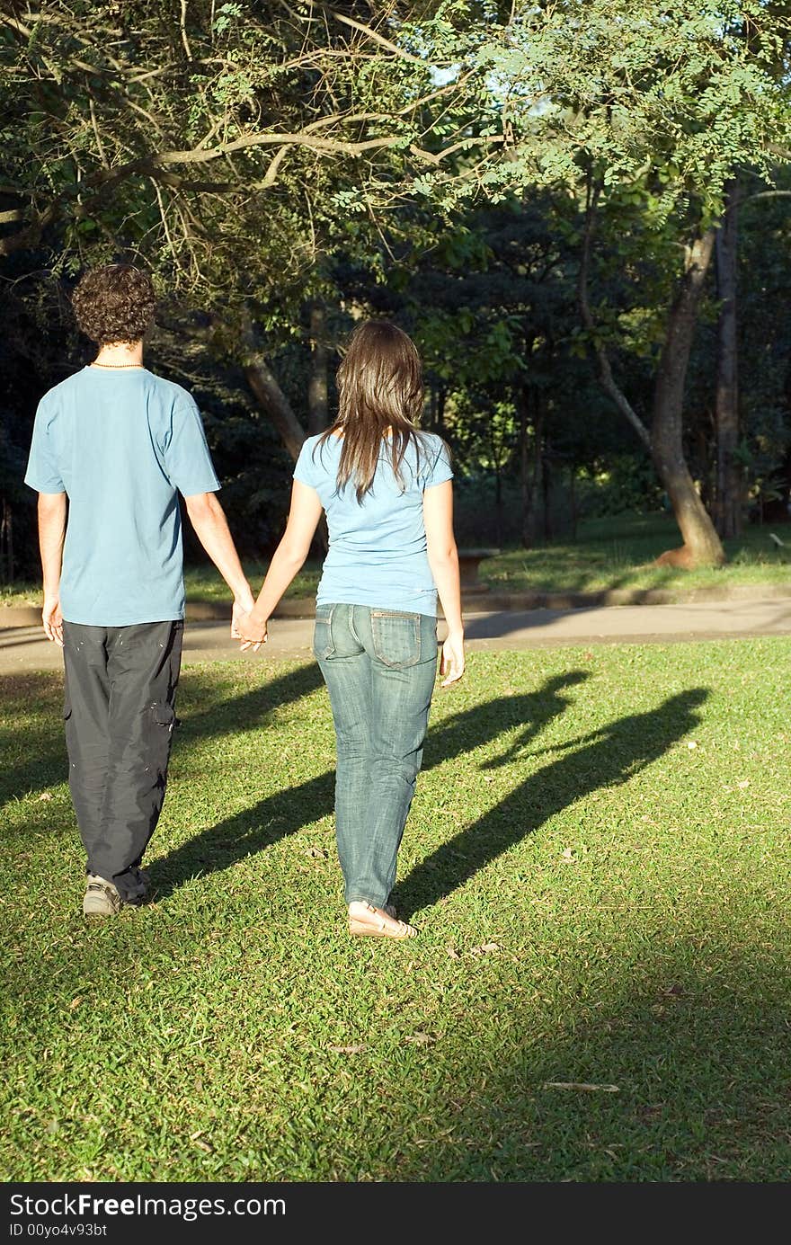 Young couple walking through a park holding hands on a sunny day. They are facing away and you can see their shadows. Vertically framed photograph. Young couple walking through a park holding hands on a sunny day. They are facing away and you can see their shadows. Vertically framed photograph