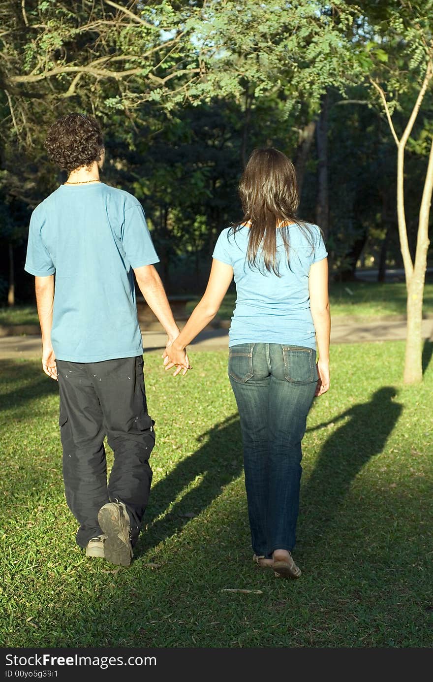 Young couple walking through a park holding hands on a sunny day. They are facing away and you can see their shadows. Vertically framed photograph. Young couple walking through a park holding hands on a sunny day. They are facing away and you can see their shadows. Vertically framed photograph