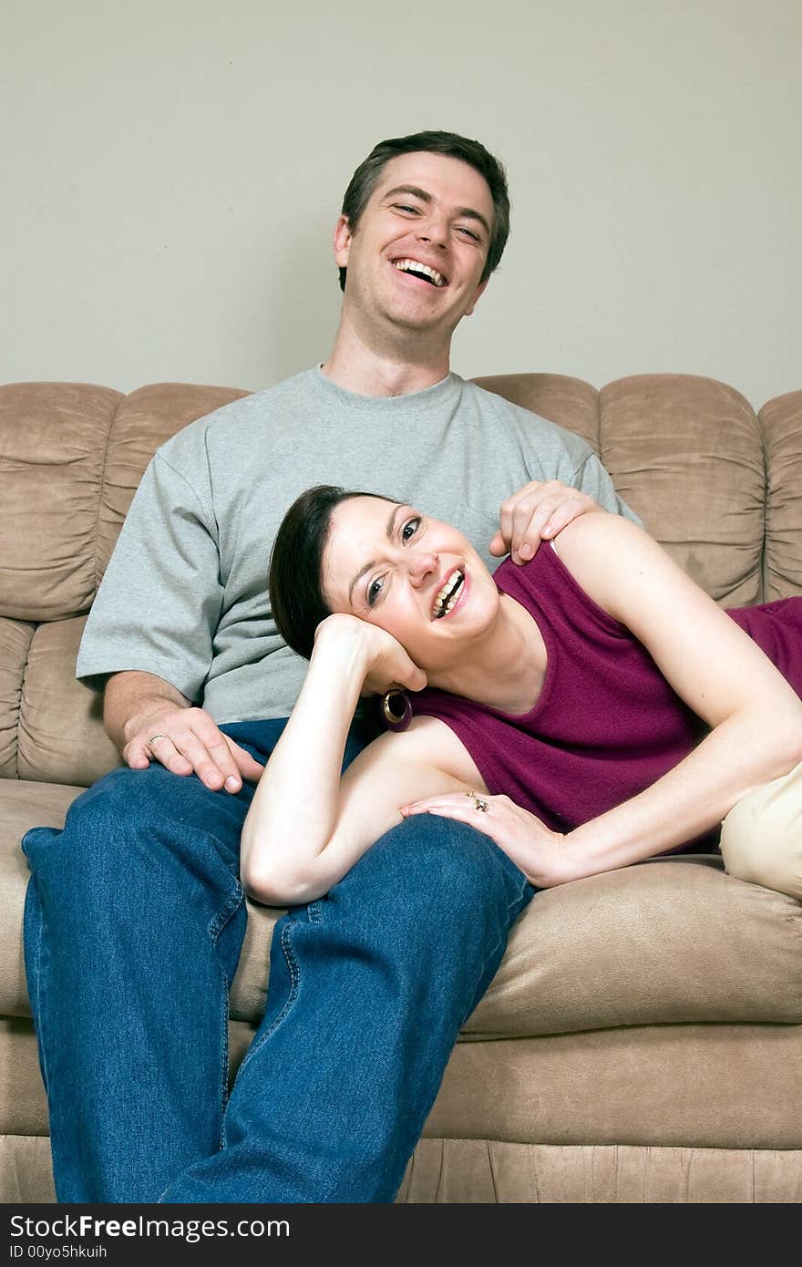 Smiling, happy couple sitting on a couch. They are laughing and he has his arm around her as she lays across his lap. Vertically framed photograph. Smiling, happy couple sitting on a couch. They are laughing and he has his arm around her as she lays across his lap. Vertically framed photograph