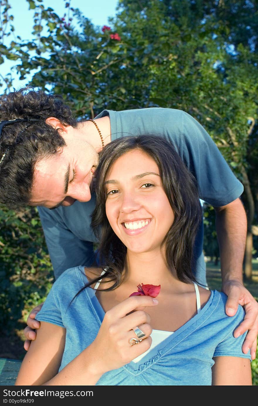 Happy couple in a park snuggle as she holds a red flower and smiles. He nuzzles her head. Vertically framed photograph. Happy couple in a park snuggle as she holds a red flower and smiles. He nuzzles her head. Vertically framed photograph.