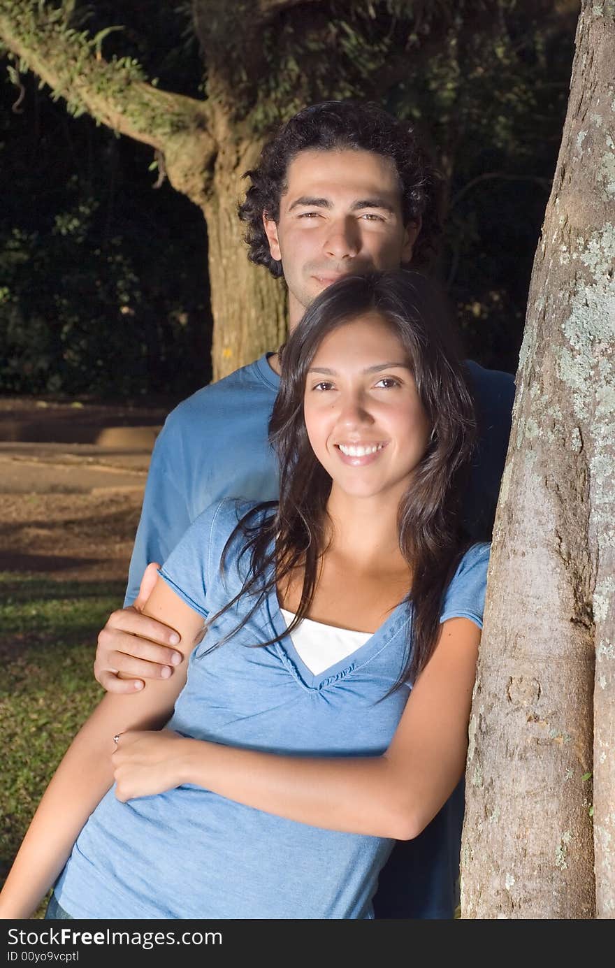 Portrait of a happy couple leaning against a tree in a park. They are smiling as he holds her arm. Vertically framed photograph. Portrait of a happy couple leaning against a tree in a park. They are smiling as he holds her arm. Vertically framed photograph.