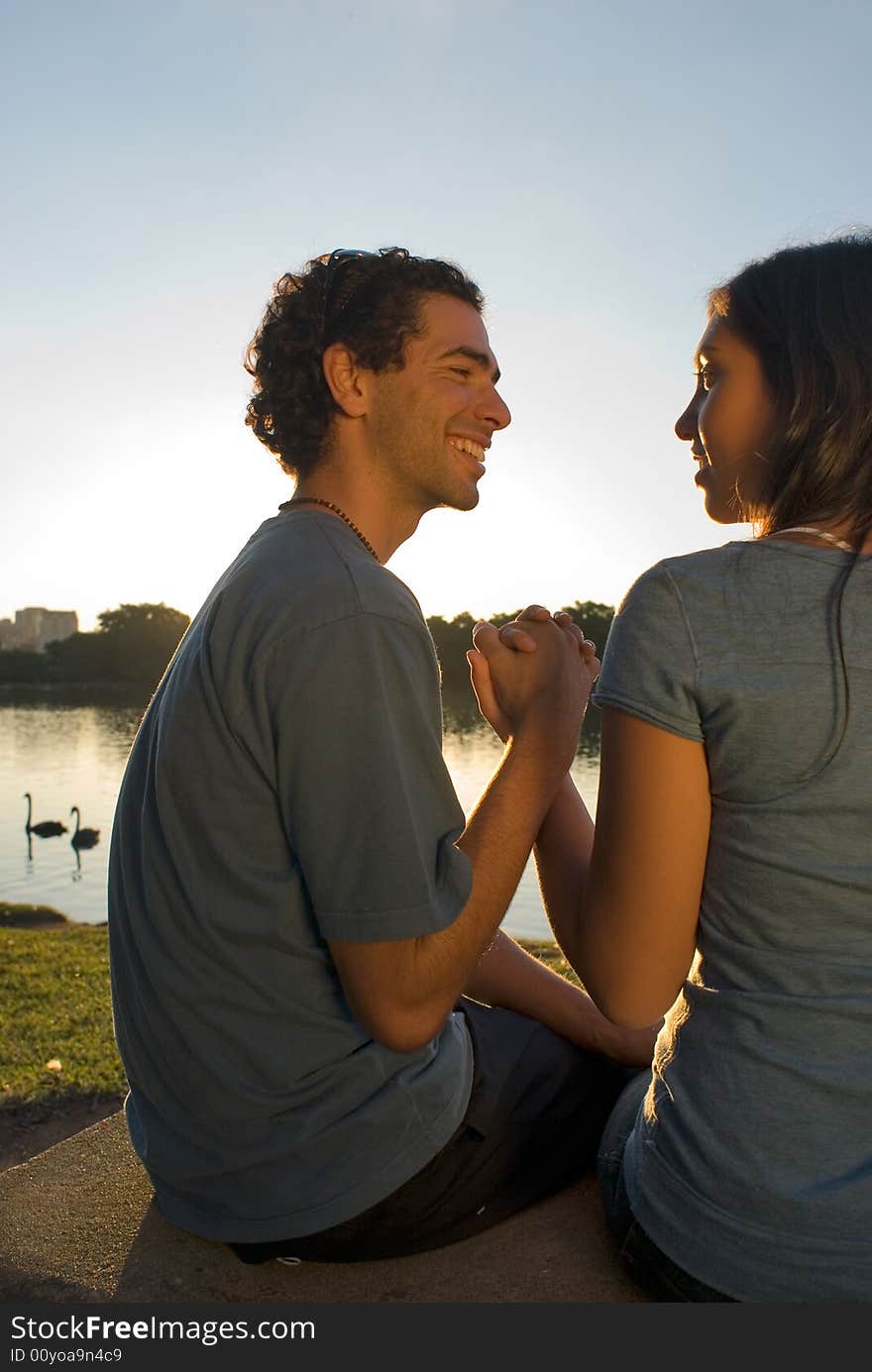 A smiling couple holding hands and gazing at each other front of a beautiful pond with swans swimming in it. Vertically framed photograph. A smiling couple holding hands and gazing at each other front of a beautiful pond with swans swimming in it. Vertically framed photograph.