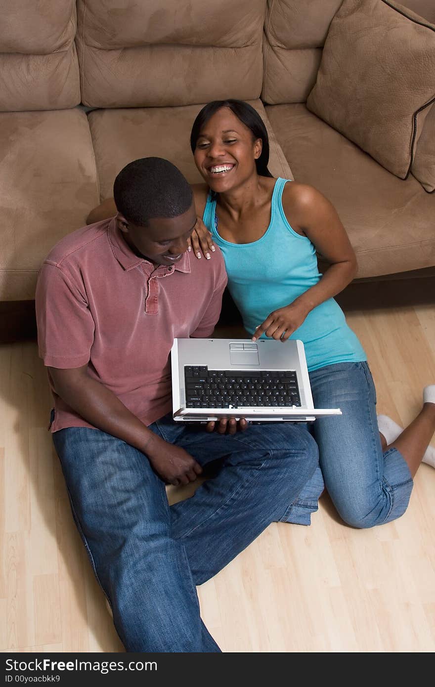 Couple sitting on the floor in front of a couch. They are looking at a laptop computer together. She has her arm on his shoulder and is laughing. Vertically framed photograph. Couple sitting on the floor in front of a couch. They are looking at a laptop computer together. She has her arm on his shoulder and is laughing. Vertically framed photograph