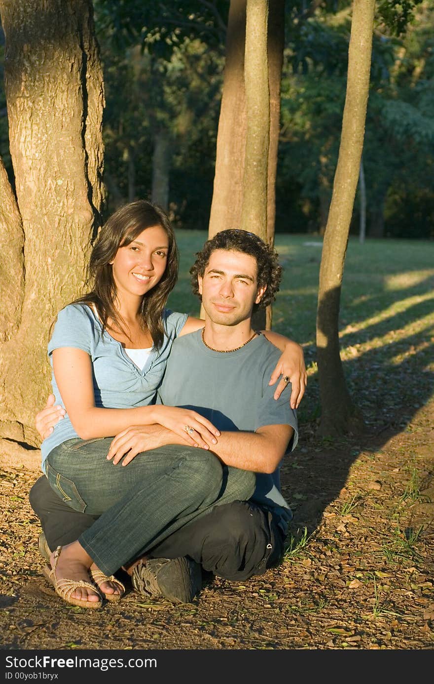 Happy couple sitting on the ground under the trees. She is sitting on his lap and they are smiling. Vertically framed photograph. Happy couple sitting on the ground under the trees. She is sitting on his lap and they are smiling. Vertically framed photograph.