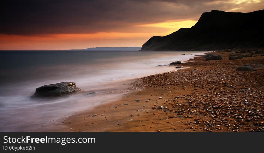 The Sun setting behind the picturesque cliffs of Eype beach in Dorset. A long exposure has resulted in soft movement in the waves as they crash on the pebble and sand beach. The Sun setting behind the picturesque cliffs of Eype beach in Dorset. A long exposure has resulted in soft movement in the waves as they crash on the pebble and sand beach