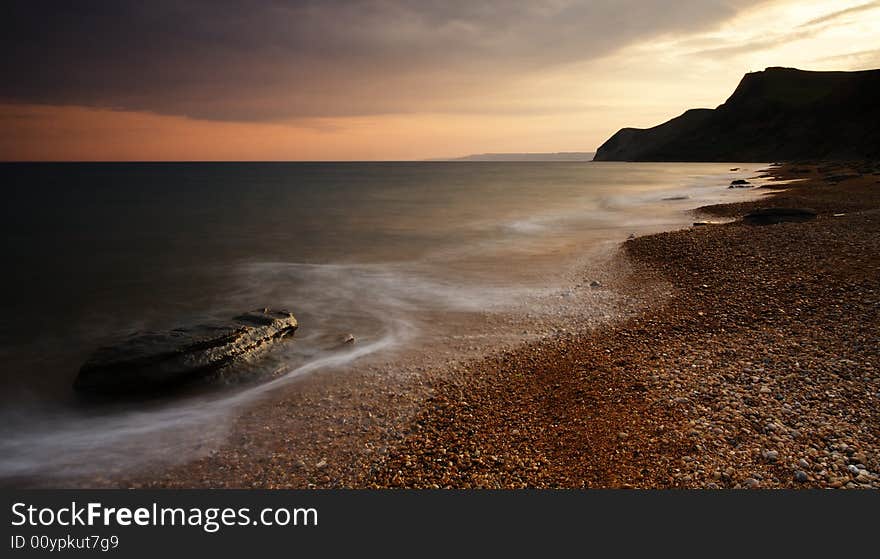 The Sun setting behind the picturesque cliffs of Eype beach in Dorset. A long exposure has resulted in soft movement in the waves as they crash on the pebble and sand beach. The Sun setting behind the picturesque cliffs of Eype beach in Dorset. A long exposure has resulted in soft movement in the waves as they crash on the pebble and sand beach