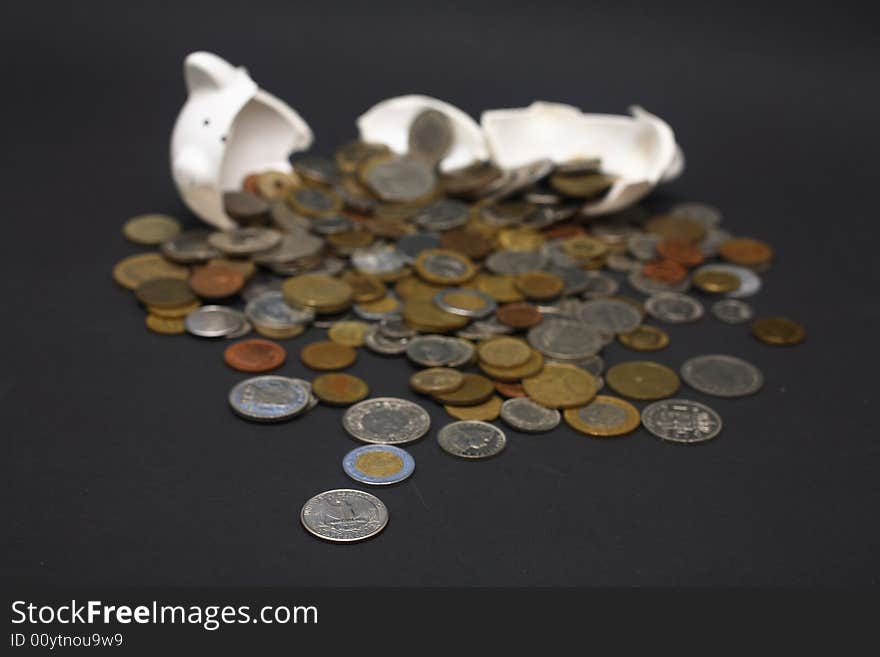 A broken piggy bank isolated on a dark background with loads of coins from around the world.