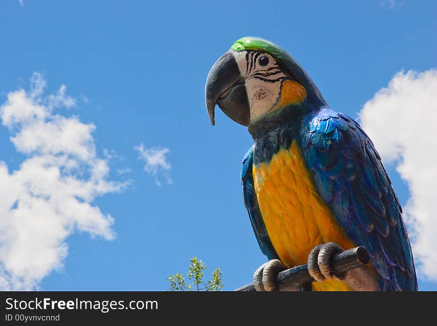 A giant stature of a blue and yellow macaw parrot on a perch with a blue sky and cloud background. Copy space to the left. A giant stature of a blue and yellow macaw parrot on a perch with a blue sky and cloud background. Copy space to the left.