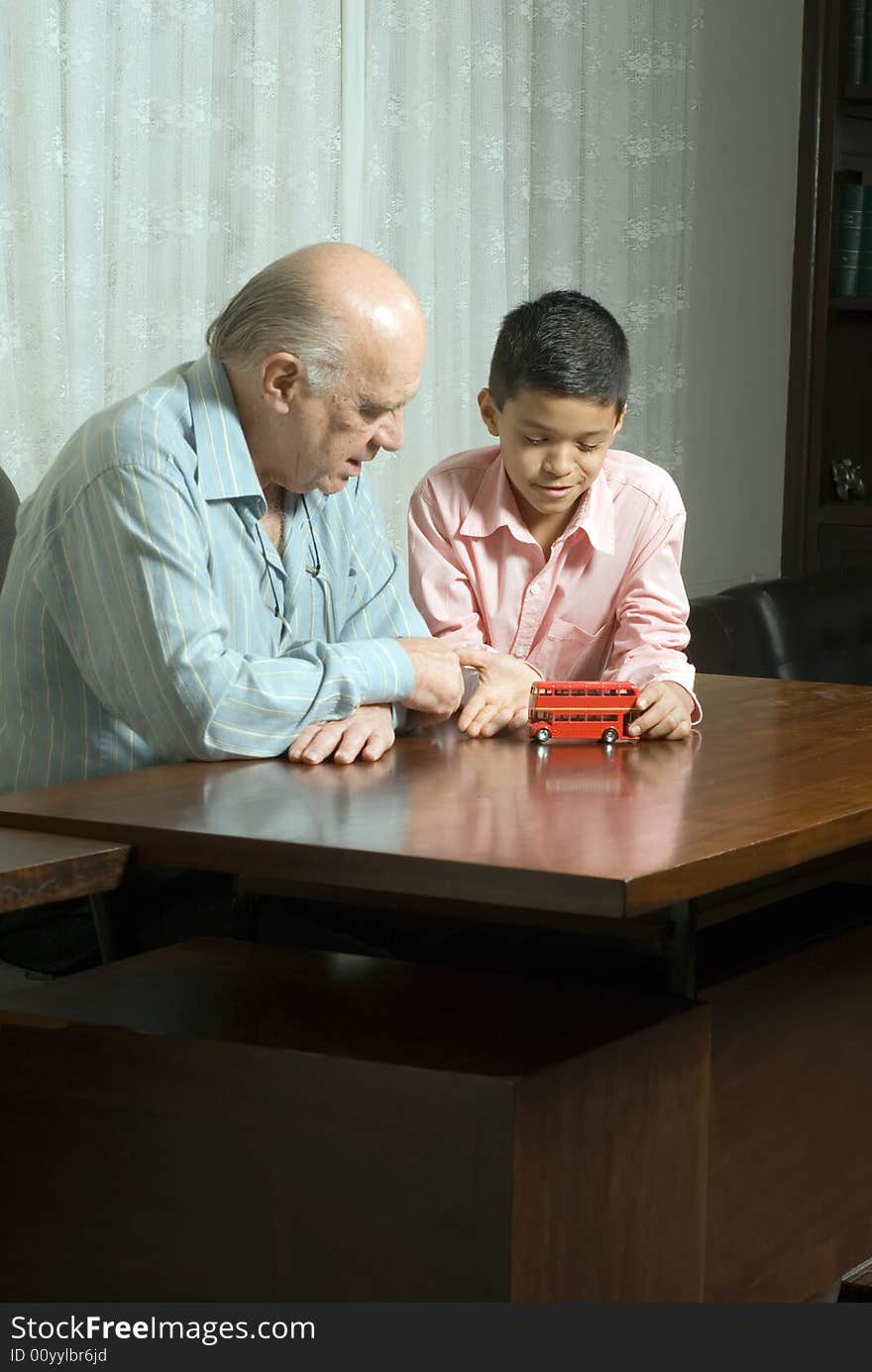 Grandfather and grandson sitting at the table with a toy bus. Grandson is playing with the bus as grandfather watches. This is a horizontally framed photo. Grandfather and grandson sitting at the table with a toy bus. Grandson is playing with the bus as grandfather watches. This is a horizontally framed photo.