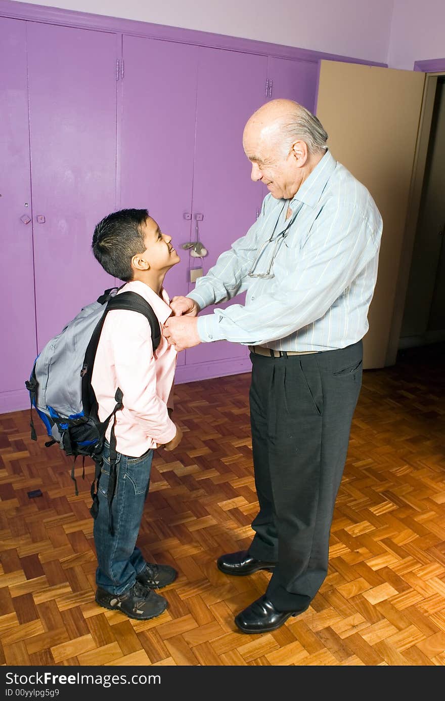 Grandfather is buttoning grandsons shirt as he is leaving for school. Grandson stands there patiently with his backpack smiling as grandfather helps him. This is a vertically framed photo. Grandfather is buttoning grandsons shirt as he is leaving for school. Grandson stands there patiently with his backpack smiling as grandfather helps him. This is a vertically framed photo.