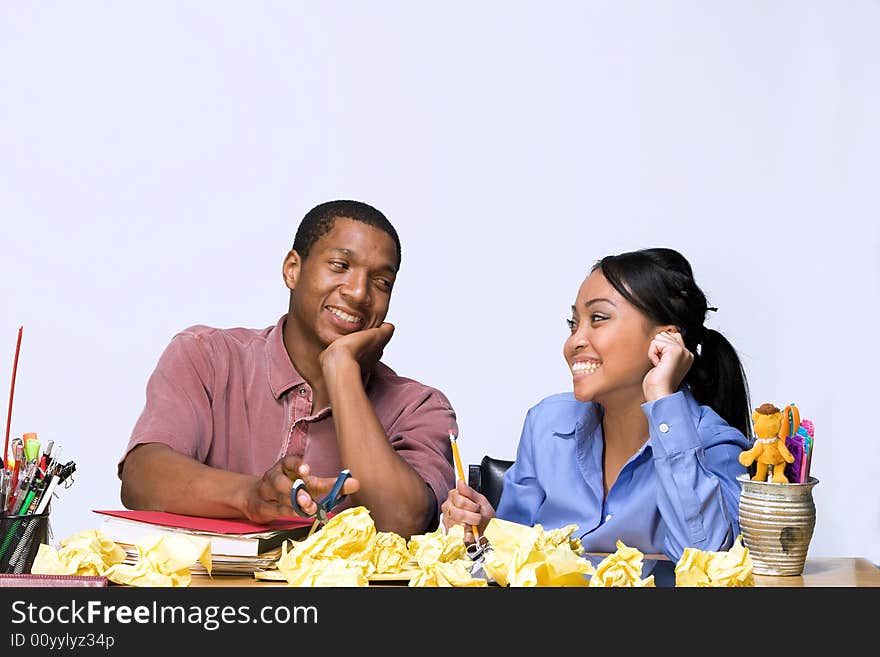 Two Teens are are seated at a desk looking at each other. There are  folders, pens, pencils, and crumpled paper on the desk. Horizontally framed photograph. Two Teens are are seated at a desk looking at each other. There are  folders, pens, pencils, and crumpled paper on the desk. Horizontally framed photograph