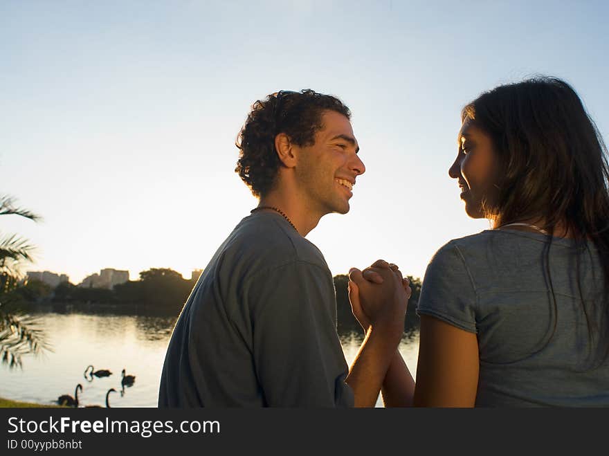 Happy couple clasp hands as they smile at each other. There is a pond with swans and buildings in the background. Horizontally framed photograph. Happy couple clasp hands as they smile at each other. There is a pond with swans and buildings in the background. Horizontally framed photograph