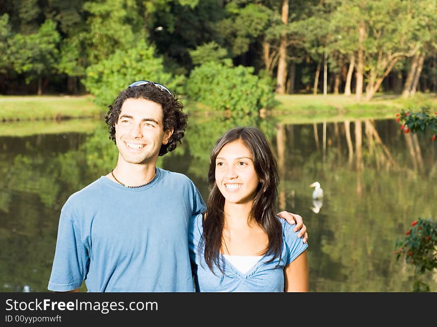 Happy couple smiling in front of a pond. He has his arm around her as they stare off in the distance. Horizontally framed photograph. Happy couple smiling in front of a pond. He has his arm around her as they stare off in the distance. Horizontally framed photograph