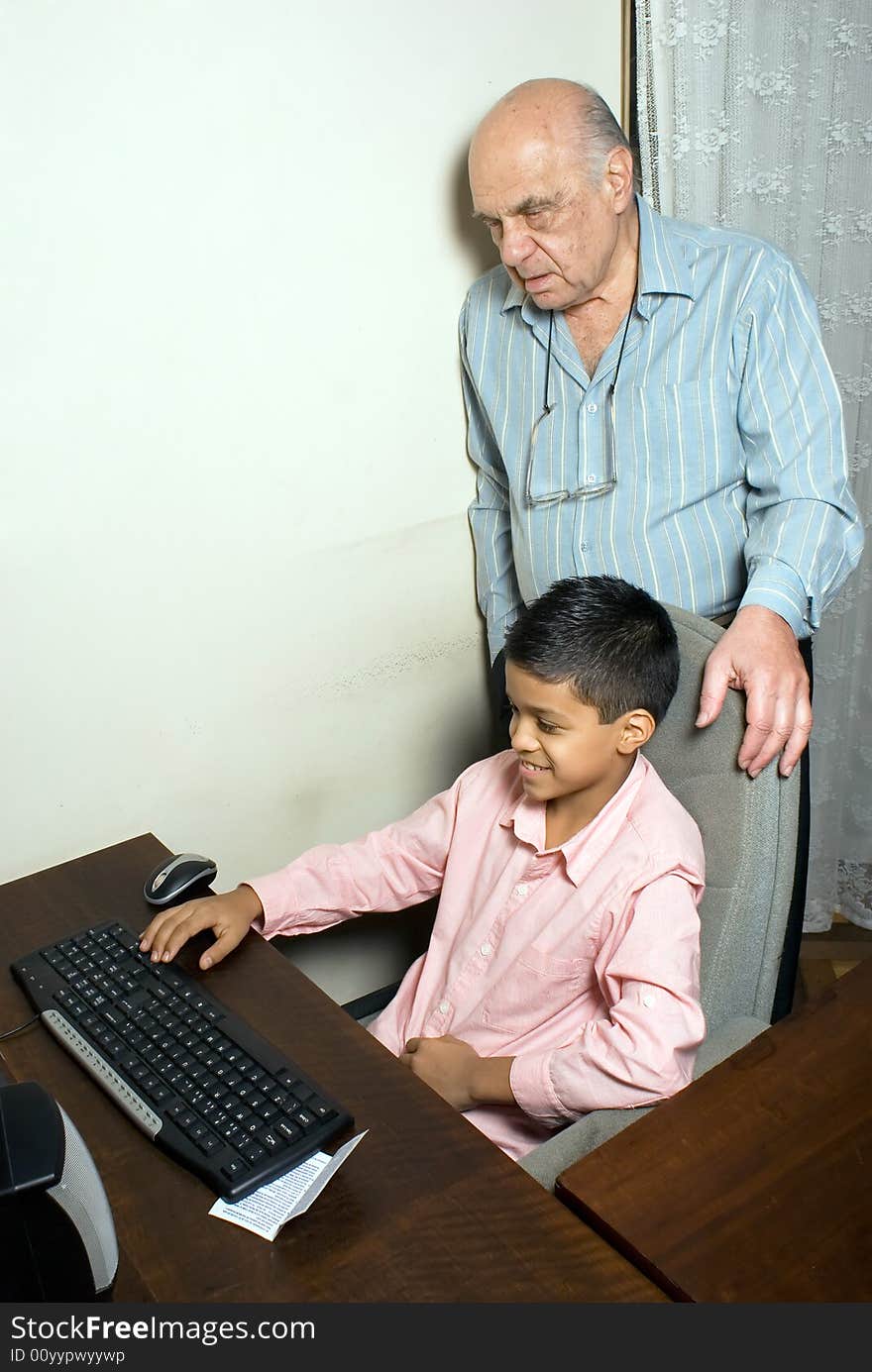 Grandfather and grandson are resting by the computer as grandfather supervises grandsons actions. Grandson uses the computer and smiles at grandfathers instructions. This is a vertically framed photo. Grandfather and grandson are resting by the computer as grandfather supervises grandsons actions. Grandson uses the computer and smiles at grandfathers instructions. This is a vertically framed photo.