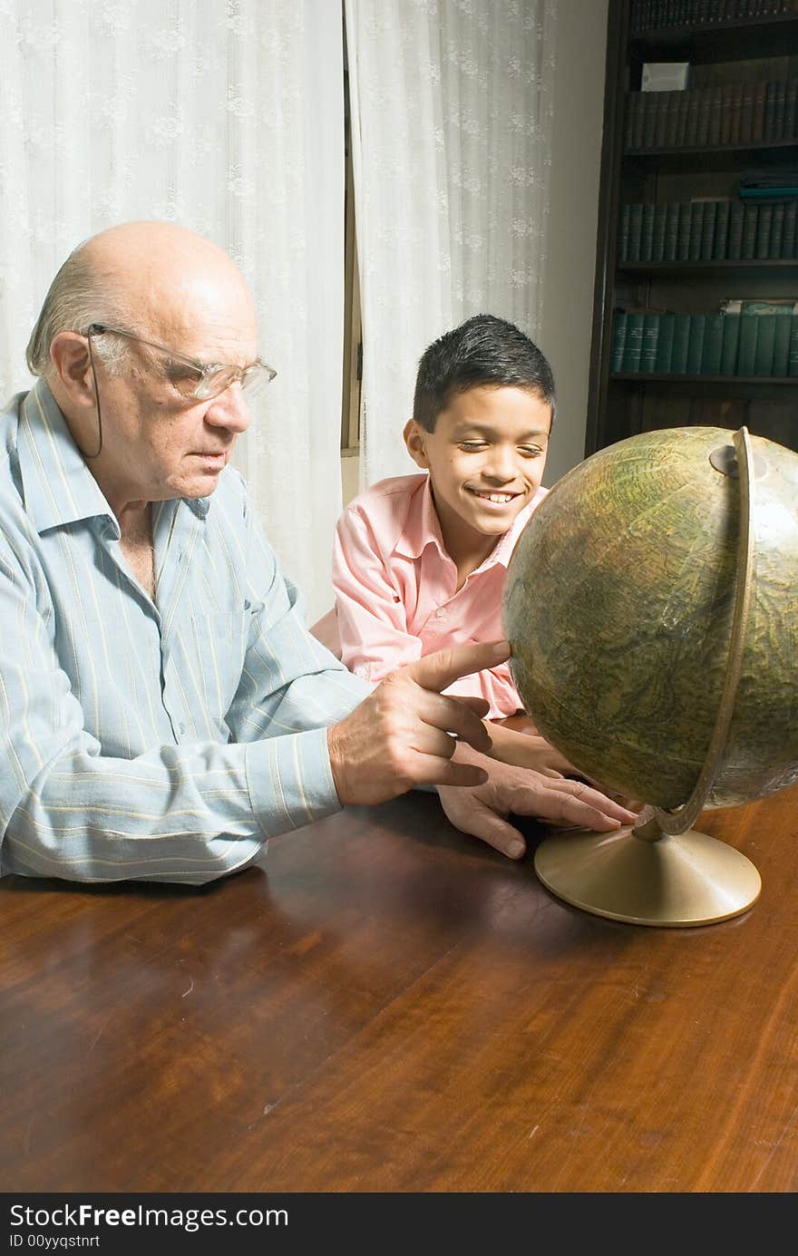 Grandfather and grandson are sitting at the table with a globe. Grandfather sits with his glasses on and points to a part of the globe as his grandson sits next to him smiling. This is a vertically framed photo. Grandfather and grandson are sitting at the table with a globe. Grandfather sits with his glasses on and points to a part of the globe as his grandson sits next to him smiling. This is a vertically framed photo.