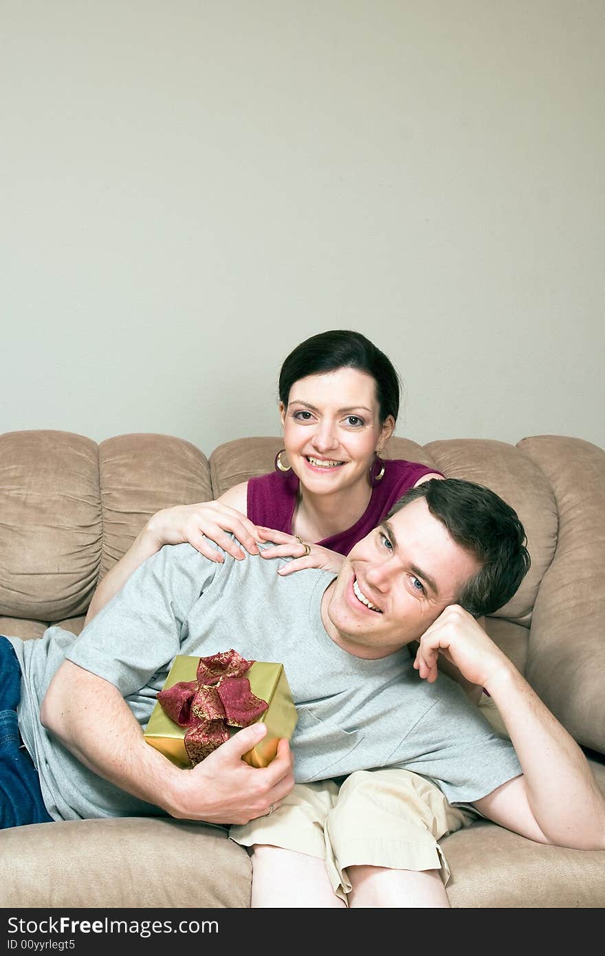 A smiling man holding a gift box lying on a smiling woman's lap on sofa. Vertically framed shot. A smiling man holding a gift box lying on a smiling woman's lap on sofa. Vertically framed shot.