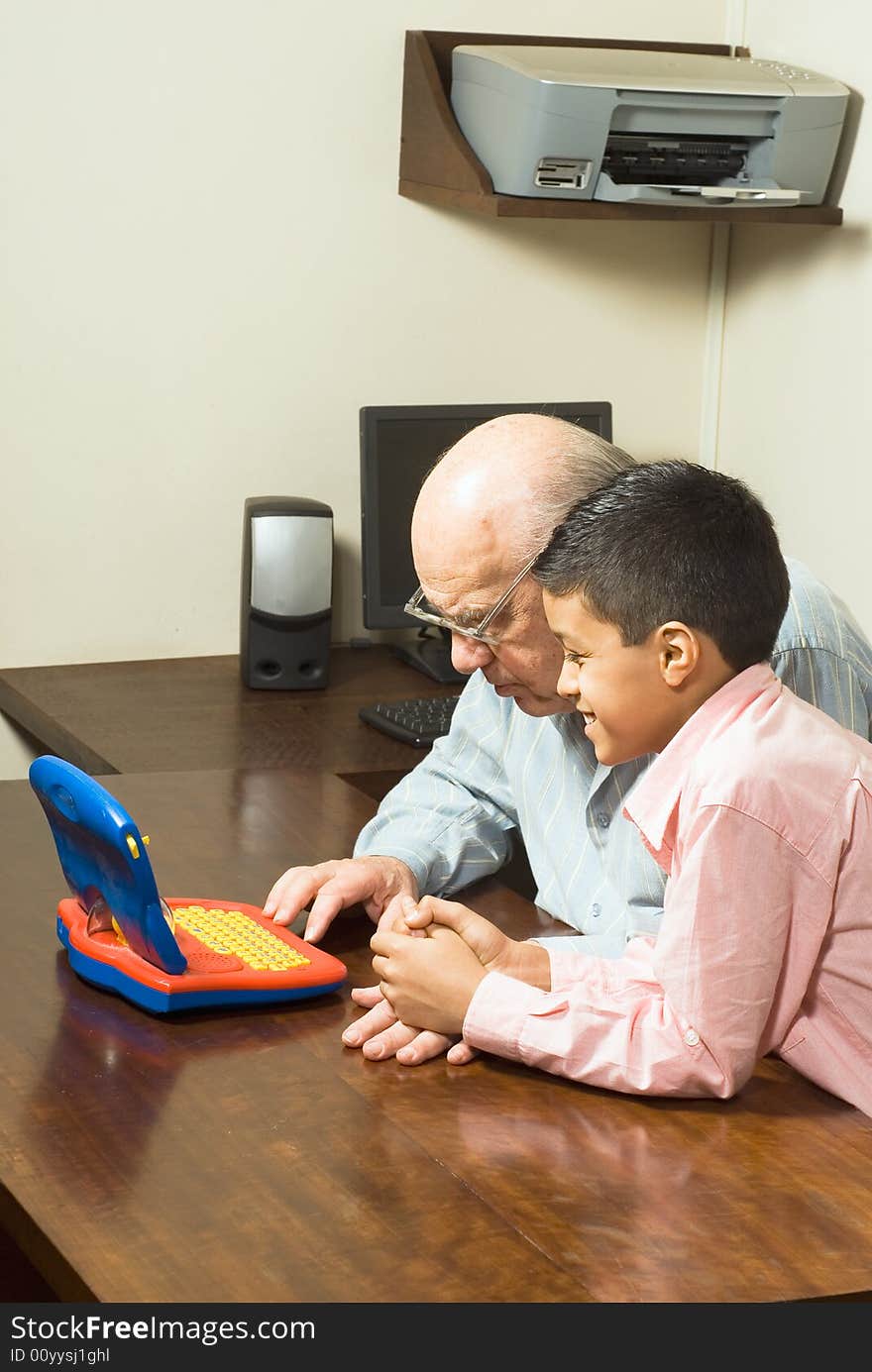 Grandfather and Grandson are seated at a table looking at a blue toy computer. There is a computer and a printer in the background. Vertically framed photograph. Grandfather and Grandson are seated at a table looking at a blue toy computer. There is a computer and a printer in the background. Vertically framed photograph.