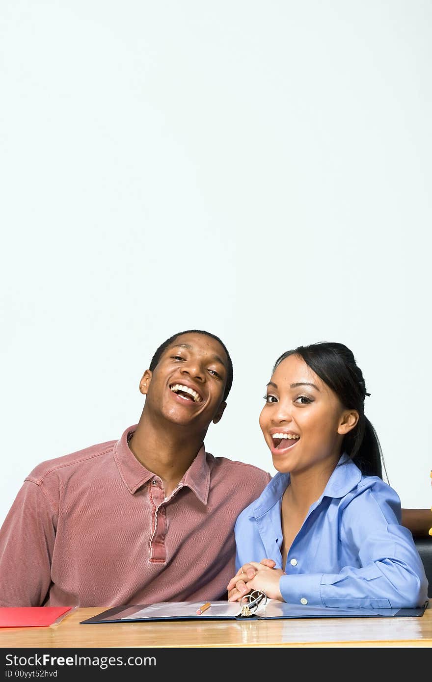 Two Teens are are seated at a desk laughing, and smiling. There are folders and paper on the desk. Vertically framed photograph. Two Teens are are seated at a desk laughing, and smiling. There are folders and paper on the desk. Vertically framed photograph