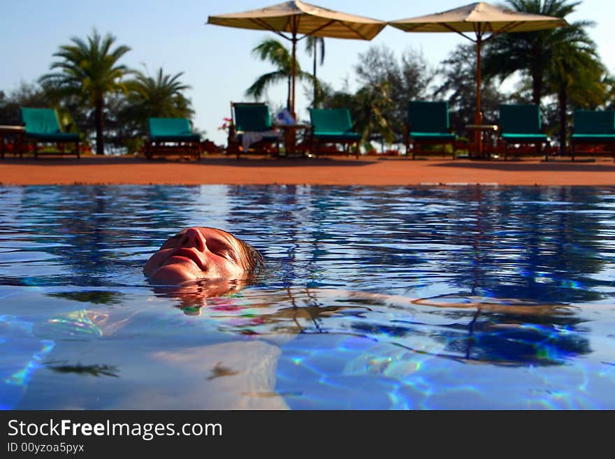 A young woman is relaxing in the pool. Ideal vacation shot with sun umbrellas and palm trees in the back.