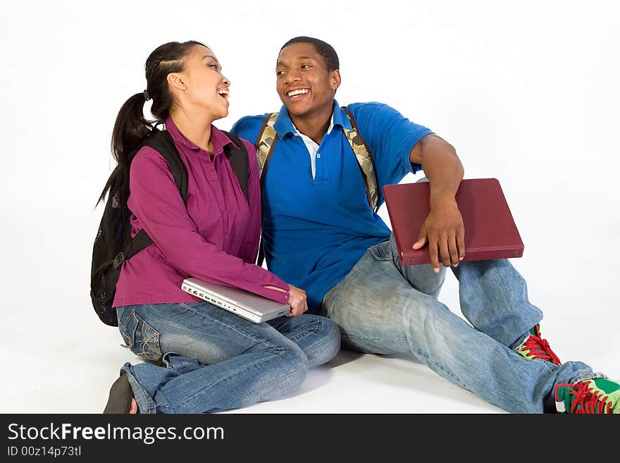 Two Students are sitting on the ground looking at each other. Both wear backpacks and he carries a notebook. Horizontally framed photograph. Two Students are sitting on the ground looking at each other. Both wear backpacks and he carries a notebook. Horizontally framed photograph