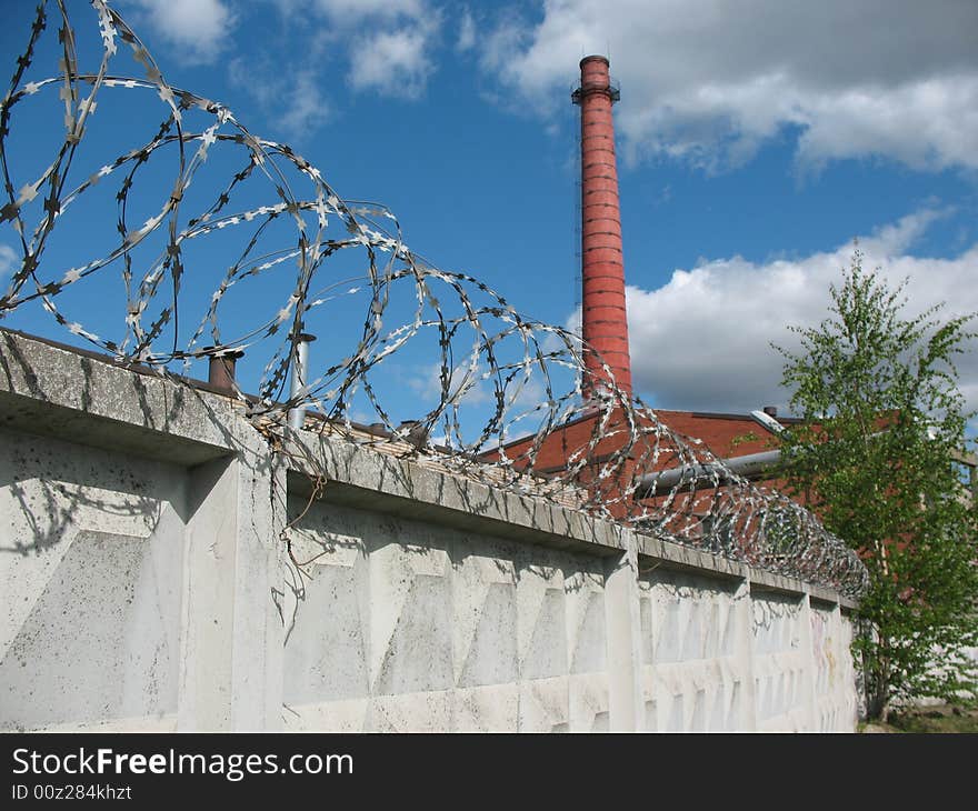 A secured industrial zone with concrete fence, barbed wire and brick chimney-stalk on the blue sky background.