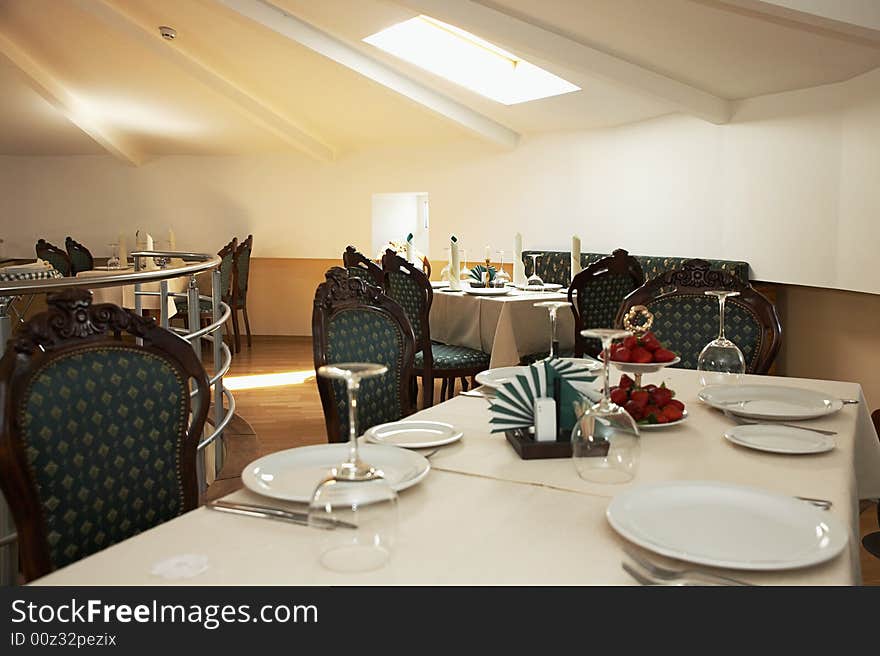 Interior of a restaurant shows laid out tables with plates, glasses, crockery and green chairs