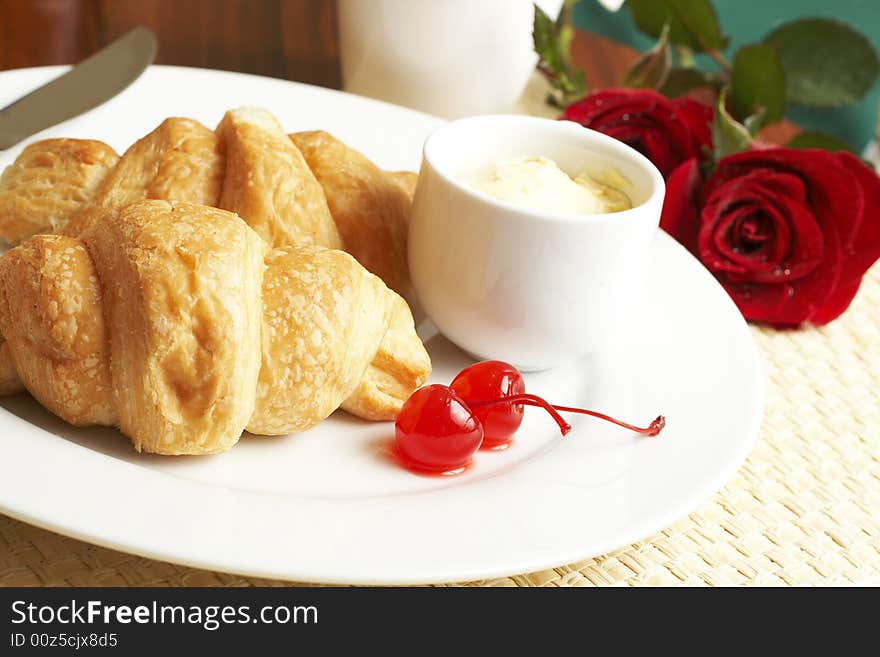 Breakfast plate with croissants, butter and glazed maraschino cherries with red roses in the background. Breakfast plate with croissants, butter and glazed maraschino cherries with red roses in the background