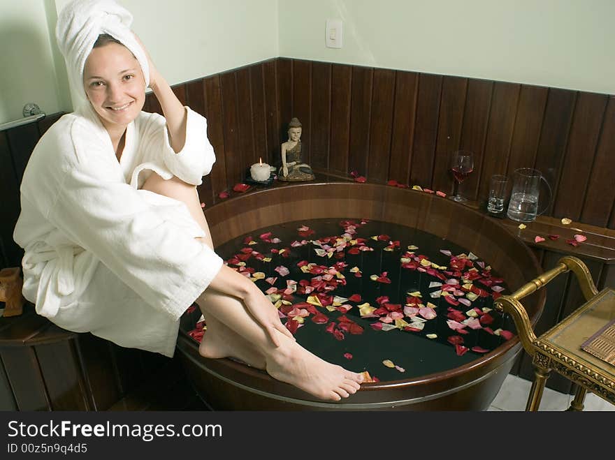 Woman in bathrobe relaxes by a spa tub filled with rose petals as she smiles. Horizontally framed photograph. Woman in bathrobe relaxes by a spa tub filled with rose petals as she smiles. Horizontally framed photograph