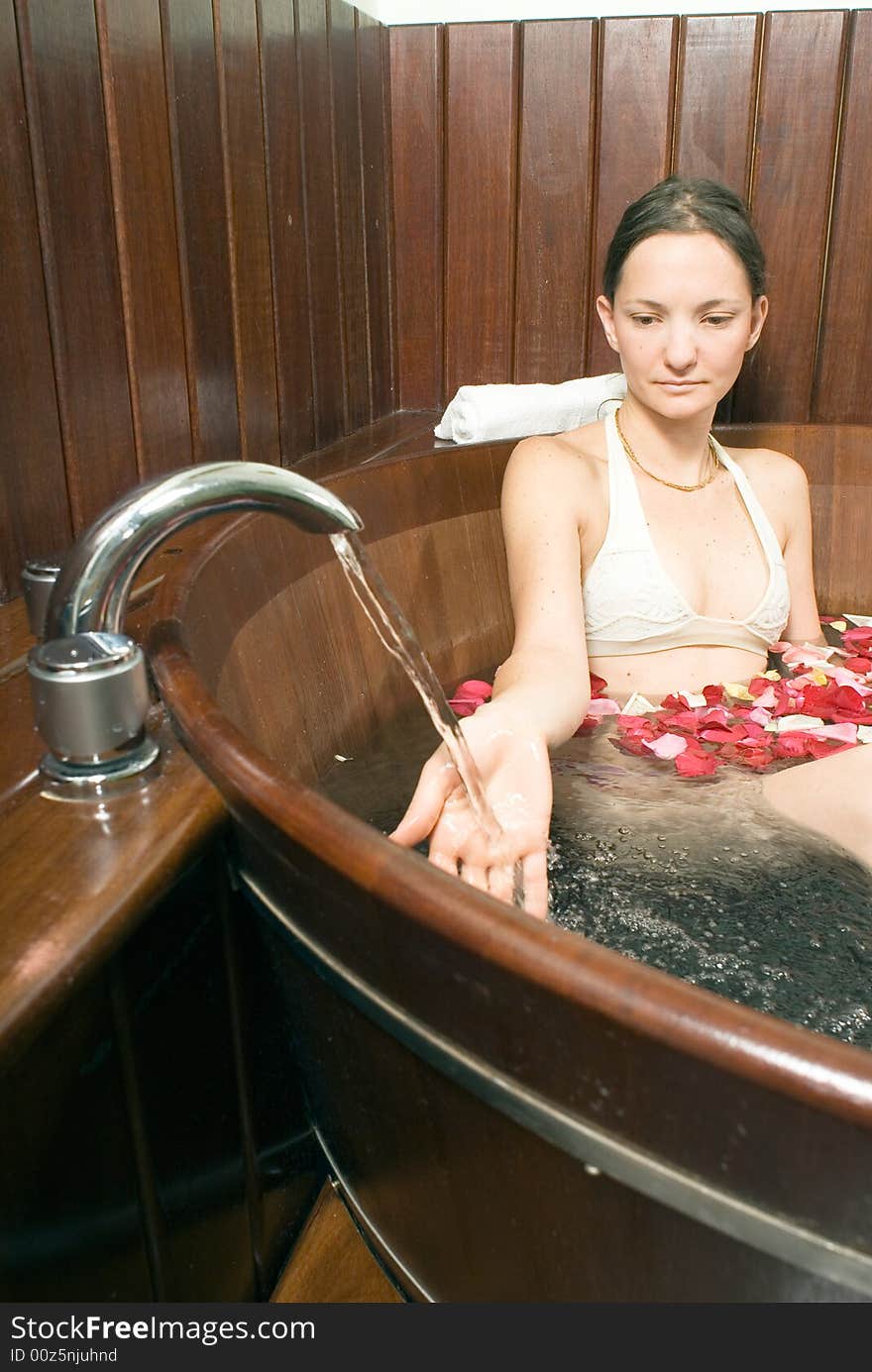 Young attractive lady, relaxing in a bathtub, playing with the running faucet. Multicolored rose petals float on the surface of the bathtub. Vertically framed shot. Young attractive lady, relaxing in a bathtub, playing with the running faucet. Multicolored rose petals float on the surface of the bathtub. Vertically framed shot.