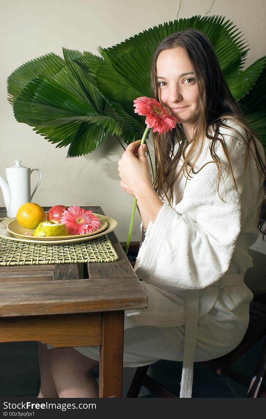 Woman smells a pink daisy as she is having fruit for breakfast. There is a large plant behind her. Vertically framed photograph. Woman smells a pink daisy as she is having fruit for breakfast. There is a large plant behind her. Vertically framed photograph.