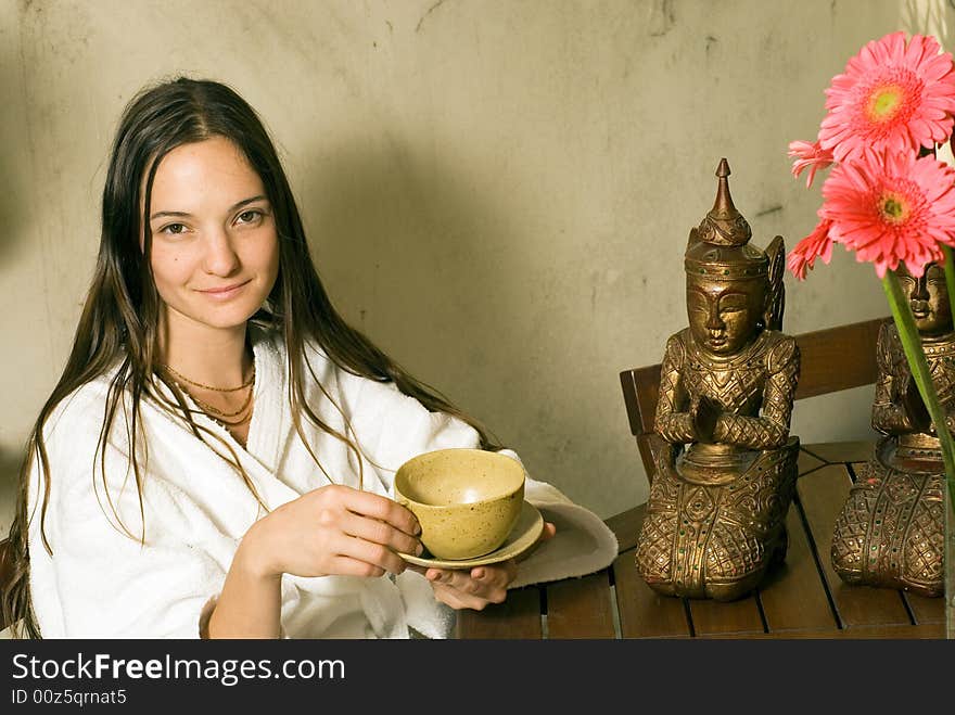 Woman smiles as she holds a tea cup. She sits next to a bronze statue and pink flowers. Horizontally framed photograph. Woman smiles as she holds a tea cup. She sits next to a bronze statue and pink flowers. Horizontally framed photograph.