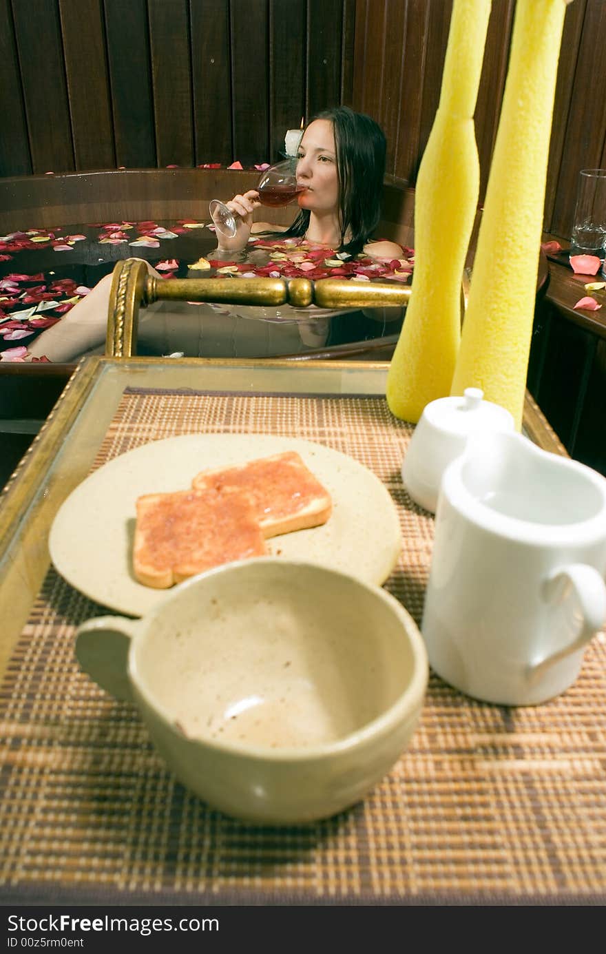 Woman relaxes in a spa tub filled with rose petals. She is drinking red wine. There are candles and lunch in the foreground. Woman relaxes in a spa tub filled with rose petals. She is drinking red wine. There are candles and lunch in the foreground.