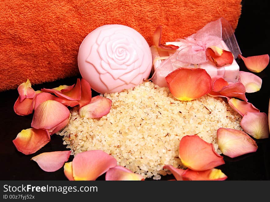 Bath salt and soap with a pink ribbon, with rose petals and bright orange towel laying on black granite. Bath salt and soap with a pink ribbon, with rose petals and bright orange towel laying on black granite