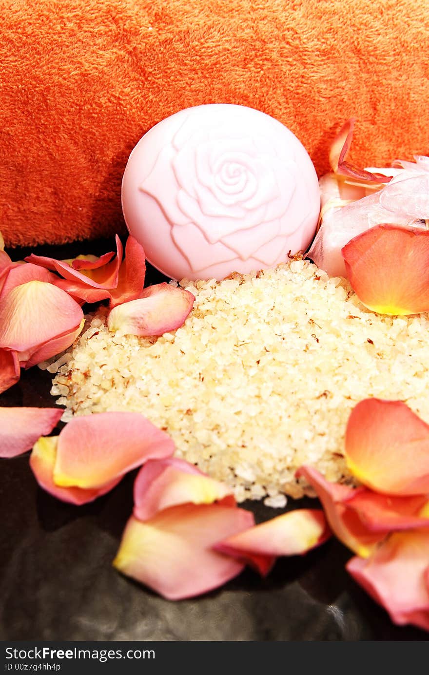 Bath salt and soap with a pink ribbon, with rose petals and bright orange towel laying on black granite. Bath salt and soap with a pink ribbon, with rose petals and bright orange towel laying on black granite