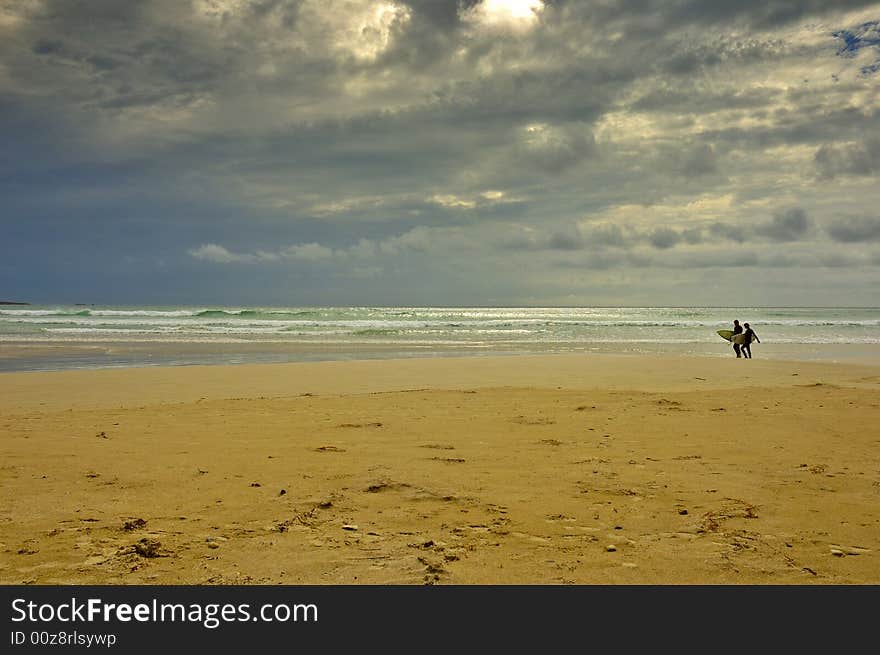 A pair of surfers walk back from the sea before the storm strikes. Space for copy in the sky. A pair of surfers walk back from the sea before the storm strikes. Space for copy in the sky.