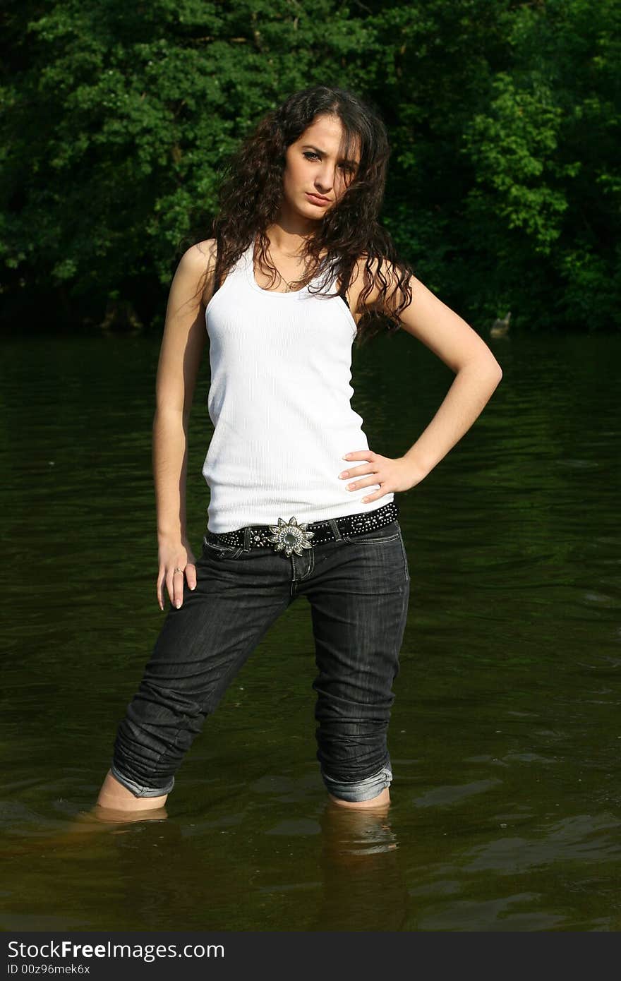 The girl in dark blue jeans and a white vest costs in water on a background of green trees. The girl in dark blue jeans and a white vest costs in water on a background of green trees