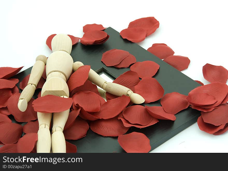 A wooden man lying on a black border mirror with red rose petal around. A wooden man lying on a black border mirror with red rose petal around