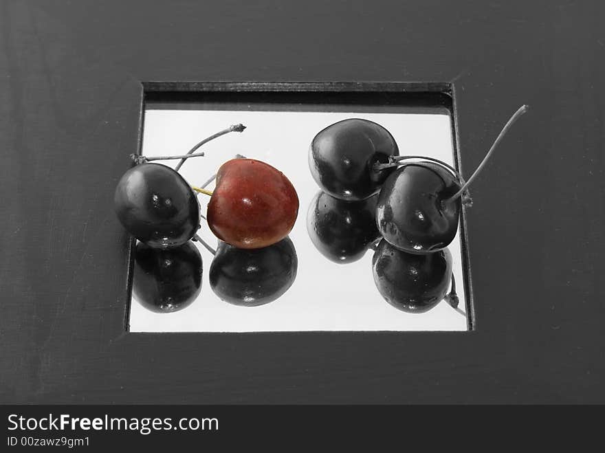 A red cherry surrounded by three black cherries on a mirror with wide black border.