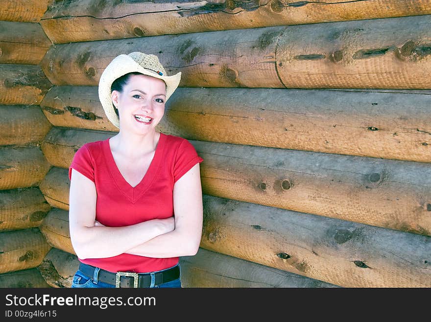 Smiling woman in cowboy hat leaning against a log cabin with her leg propped up. Horizontally framed photograph. Smiling woman in cowboy hat leaning against a log cabin with her leg propped up. Horizontally framed photograph