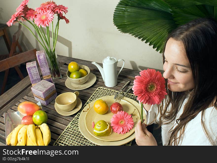 An attractive young girl outside, smelling a pink flower, sitting at a table, while fruits and plants surround her. - horizontally framed. An attractive young girl outside, smelling a pink flower, sitting at a table, while fruits and plants surround her. - horizontally framed