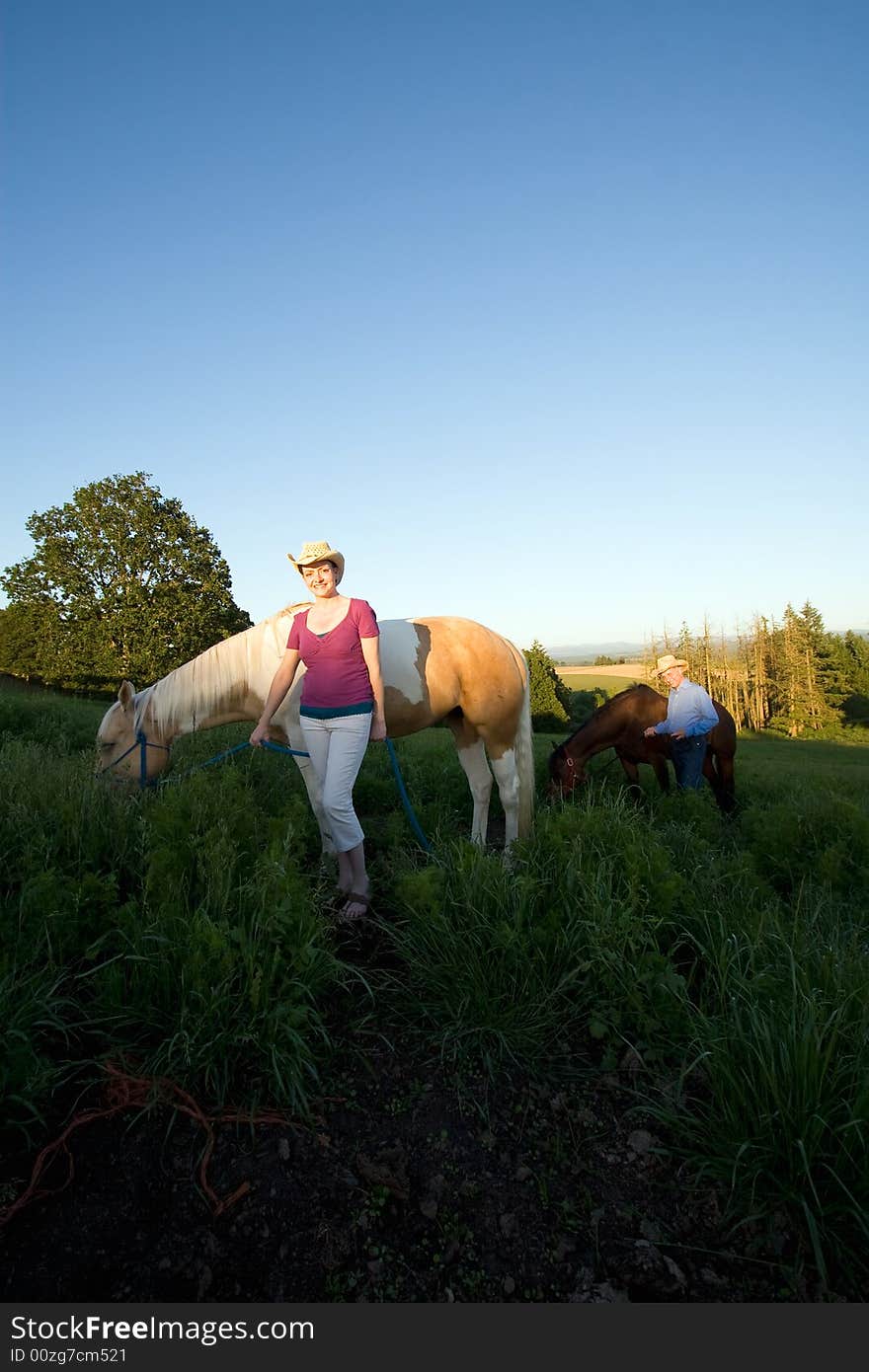 A female farmer smiles at the camera, while walking, with her horse by her side, standing in grass. Another farmer and horse are behind them. - vertically framed. A female farmer smiles at the camera, while walking, with her horse by her side, standing in grass. Another farmer and horse are behind them. - vertically framed