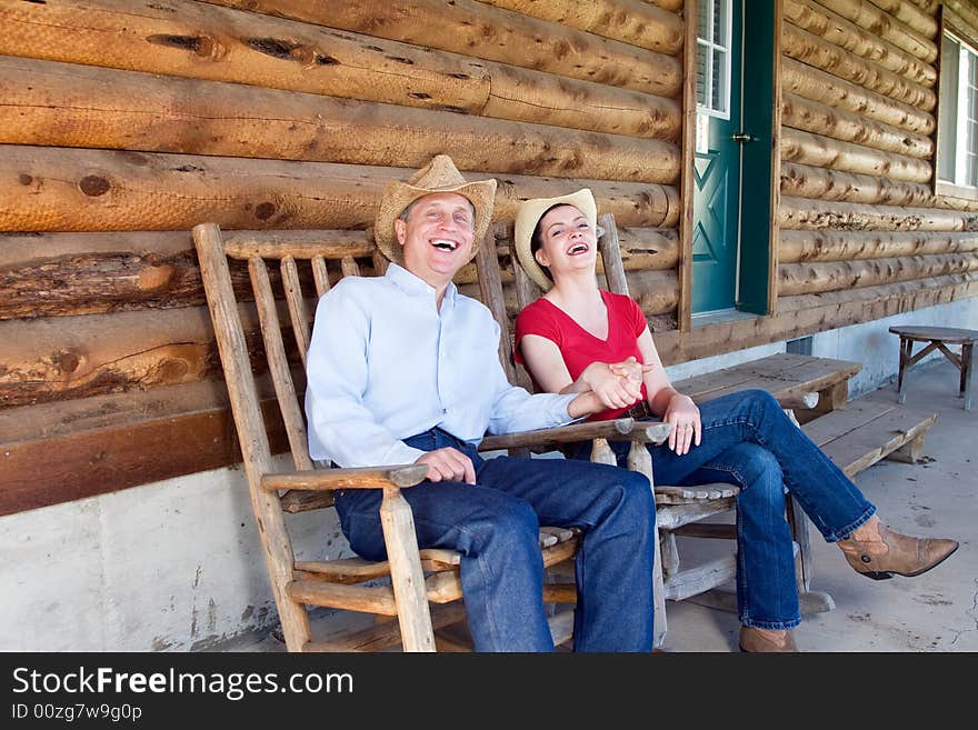 A Cowgirl and cowboy laughing and holding hands as they sit together on a porch.  Horizontally framed photograph. A Cowgirl and cowboy laughing and holding hands as they sit together on a porch.  Horizontally framed photograph