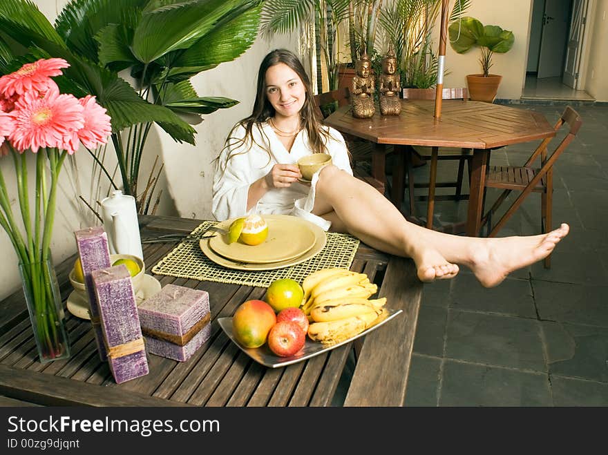 A young girl sitting in a chair, in a white bathrobe, holding a cup, surrounded by fruits and plants. - horizontally framed. A young girl sitting in a chair, in a white bathrobe, holding a cup, surrounded by fruits and plants. - horizontally framed