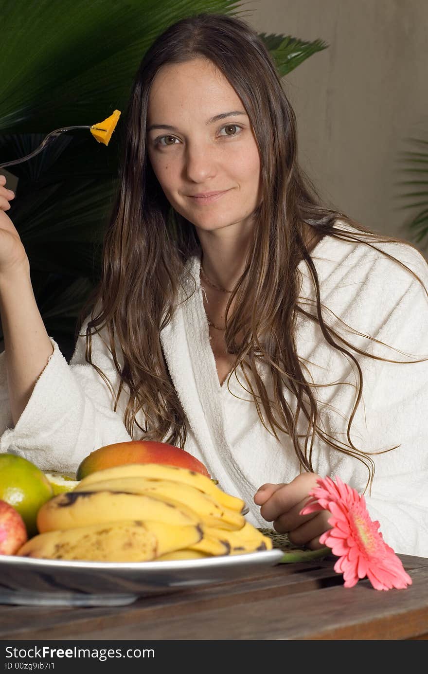 A young girl, sitting at a table full of fruits in a white bathrobe eating a mango. - vertically framed. A young girl, sitting at a table full of fruits in a white bathrobe eating a mango. - vertically framed